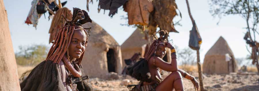 Himba-Stammesfrauen zeigt, die vor ihren Hütten in einem traditionellen Himba-Dorf in der Nähe von Kamanjab im Norden Namibias