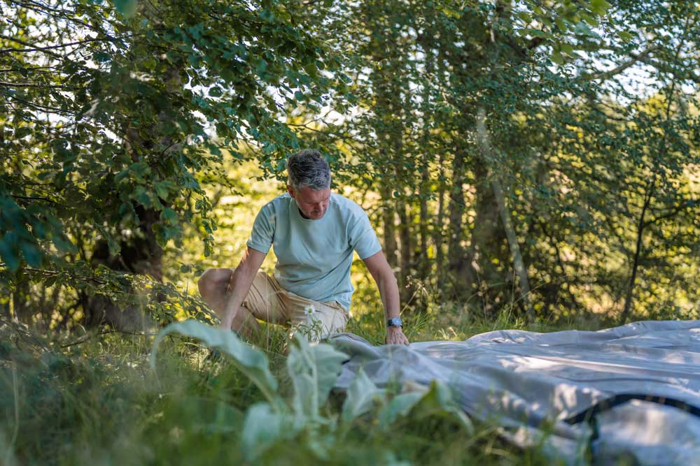 Mann breitet beim Zeltaufbau die Zeltplane auf einer Wiese am Waldrand aus